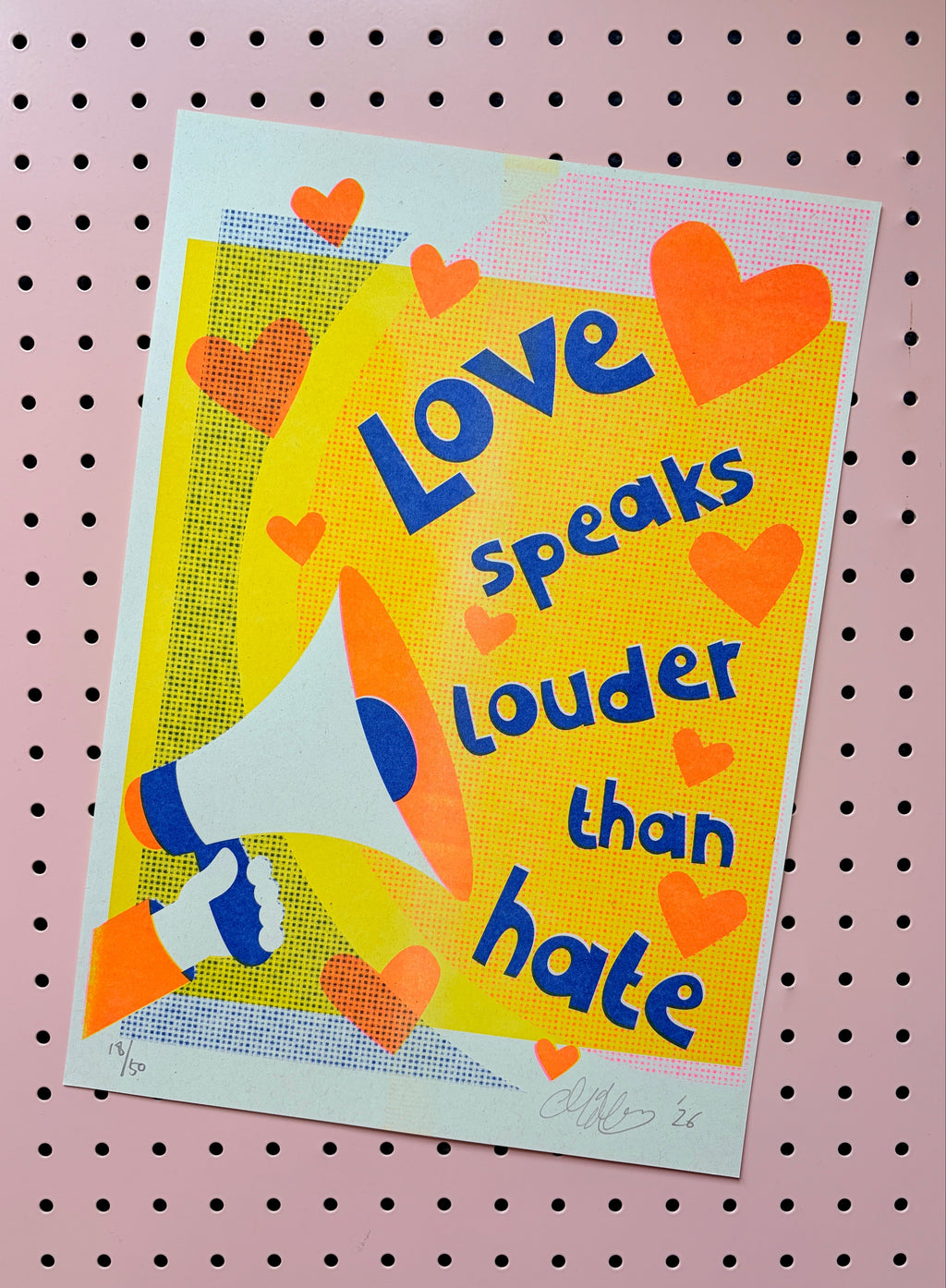 A square art print featuring the quote 'Love speaks louder than hate' in blue text over a bright yellow background, decorated with orange hearts and an illustration of a person holding a megaphone. The print is displayed on a pink pegboard background.