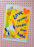 A square art print featuring the quote 'Love speaks louder than hate' in blue text over a bright yellow background, decorated with orange hearts and an illustration of a person holding a megaphone. The print is displayed on a pink pegboard background.