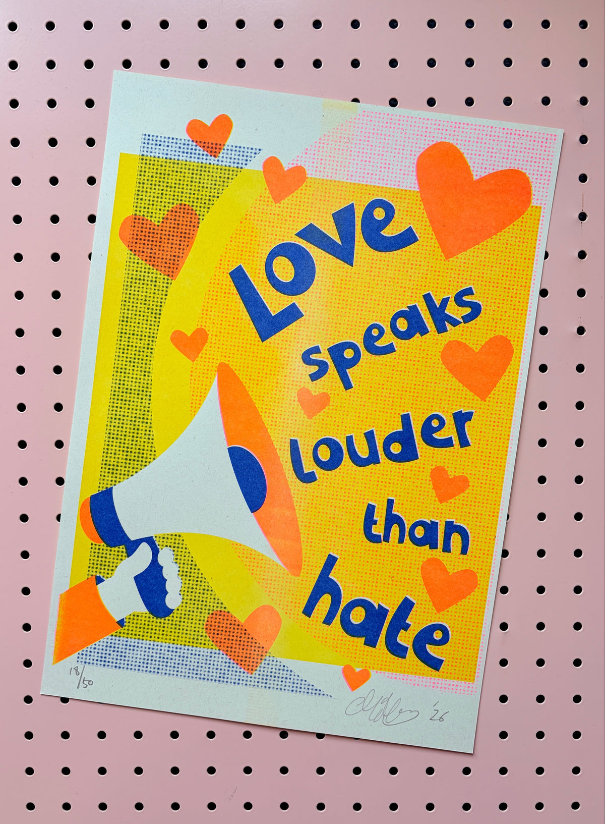 A square art print featuring the quote 'Love speaks louder than hate' in blue text over a bright yellow background, decorated with orange hearts and an illustration of a person holding a megaphone. The print is displayed on a pink pegboard background.